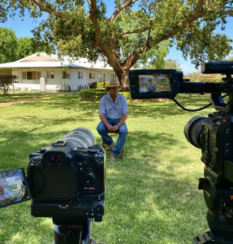 Cameras filming a seated interview on the lawn outside a rural homestead