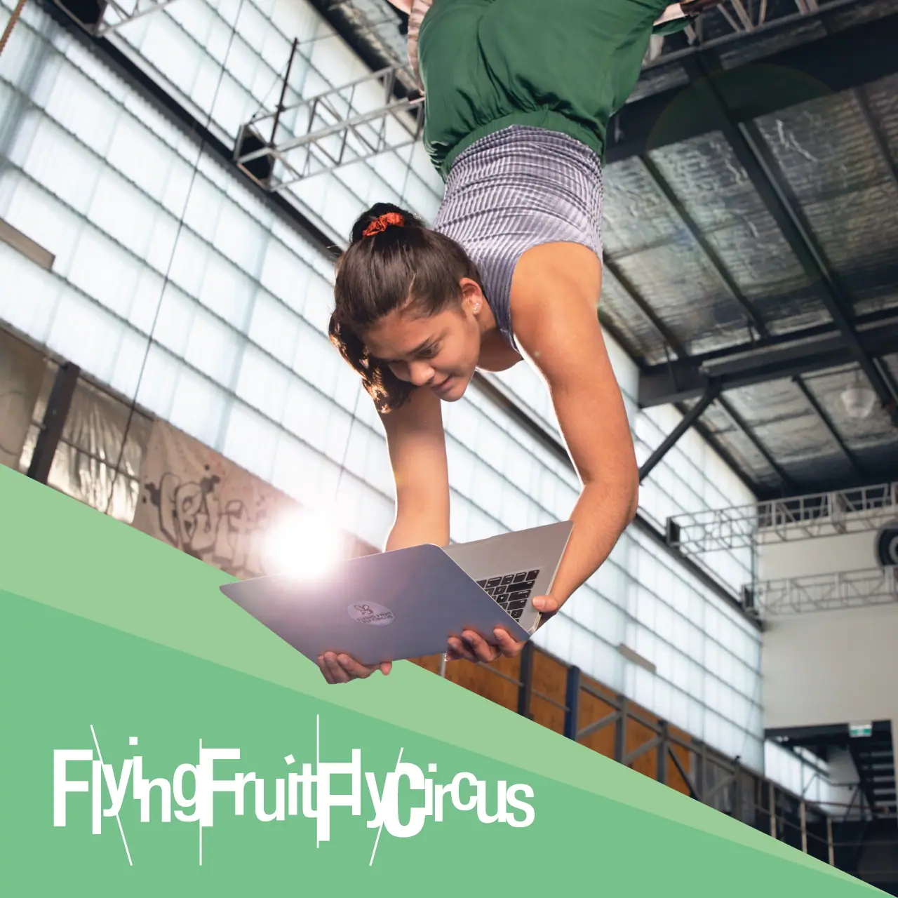 Flying Fruit Fly Circus brand graphic over a young performer using a laptop in a training hall