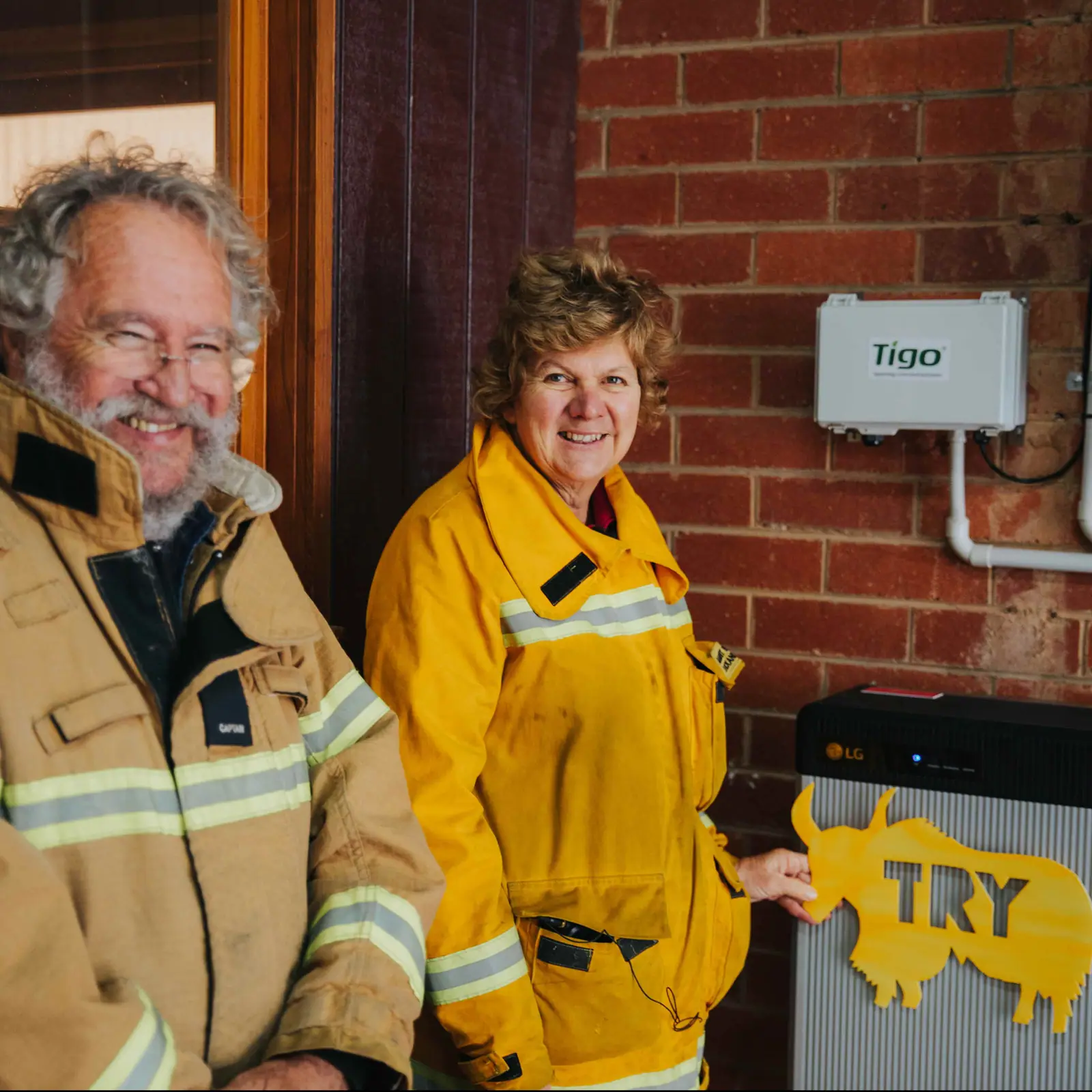 Two people in yellow firefighter jackets standing beside a Tigo solar controller and LG battery