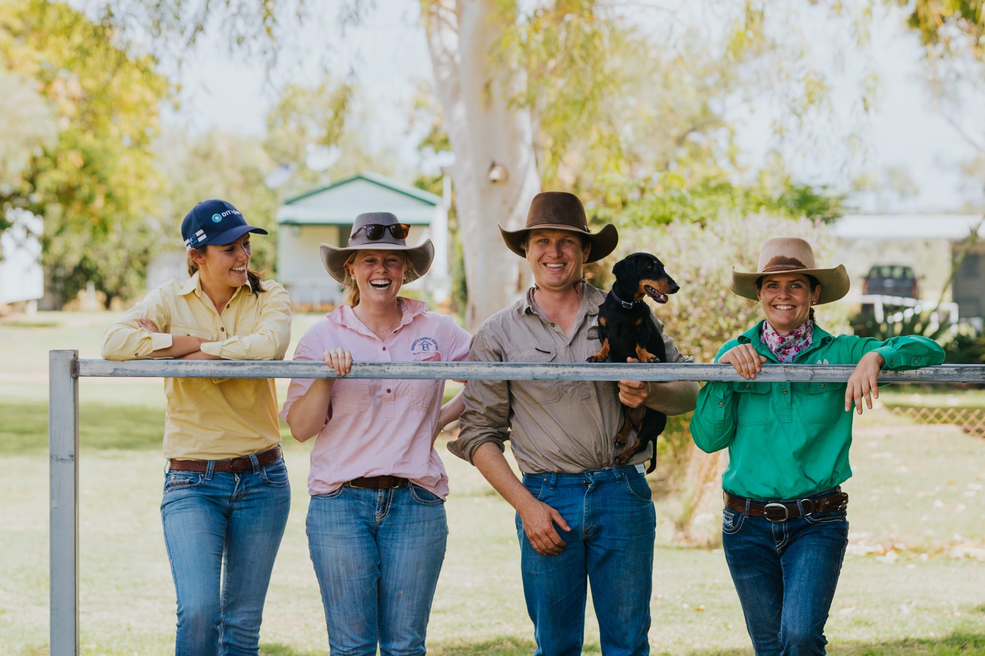 Four producers in hats leaning on a farm gate with a small black dog