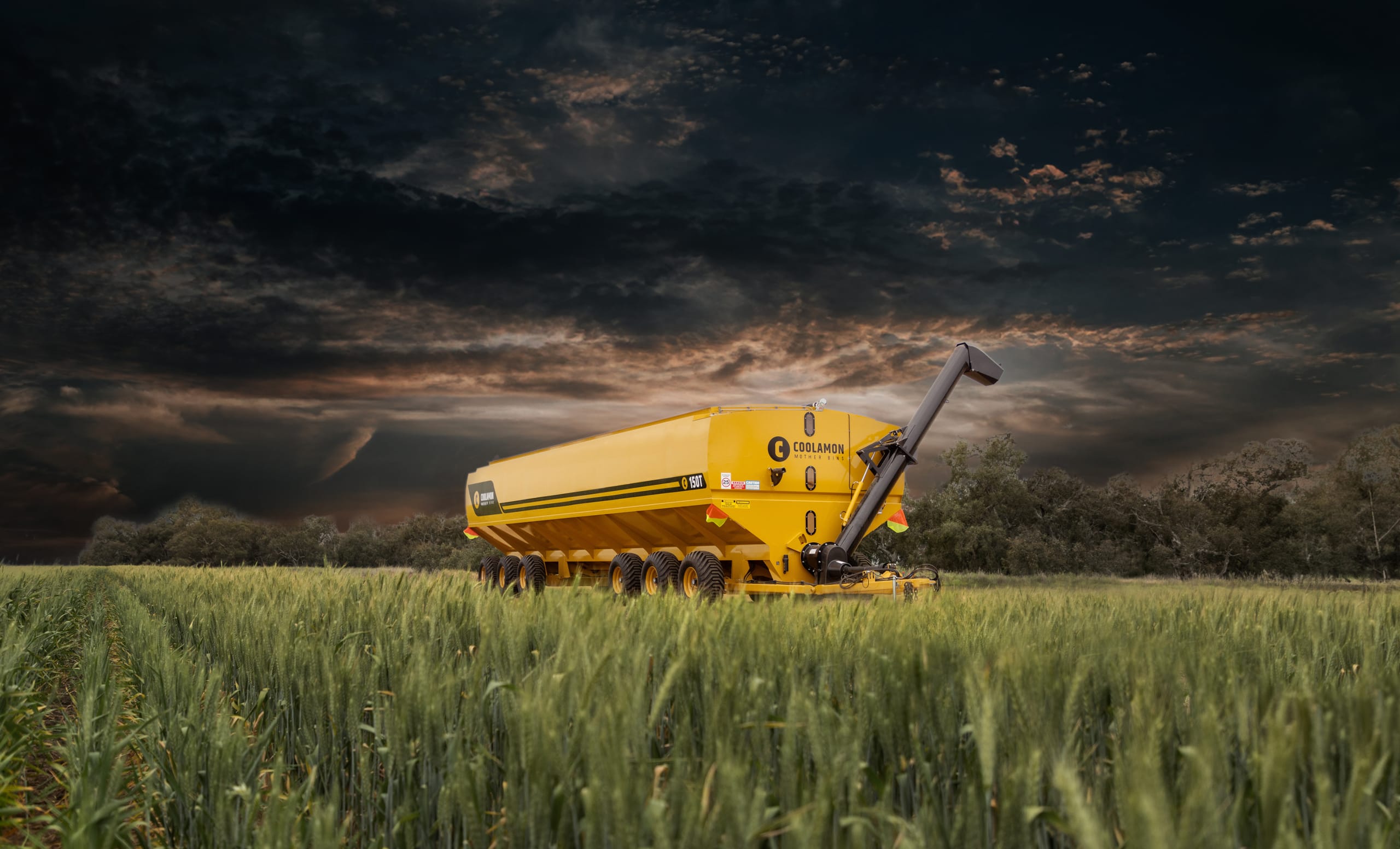 Yellow Coolamon Mother Bins grain cart in a crop field beneath a dramatic stormy sky