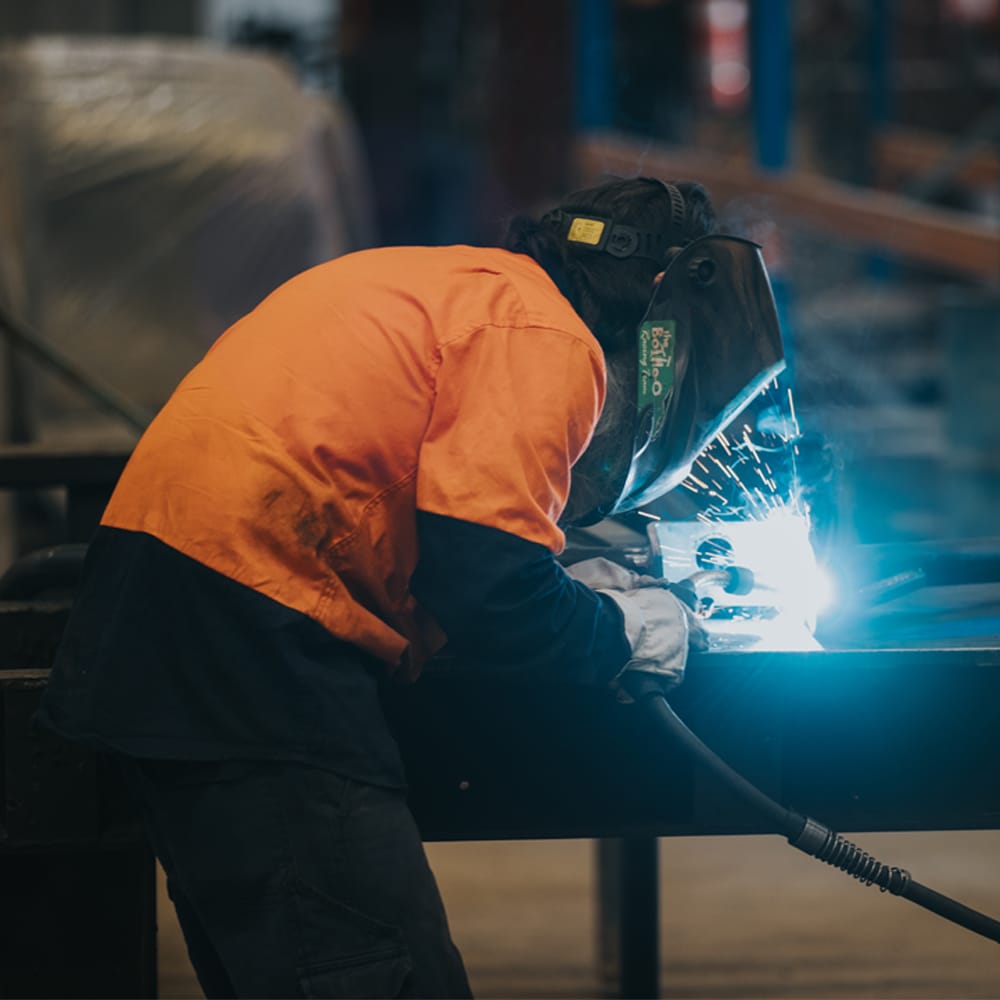 Welder in an orange high-visibility shirt fabricating a metal frame with bright welding sparks