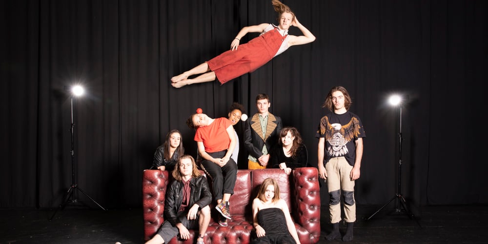Young circus ensemble posing onstage around a red sofa while one performer appears to float above them