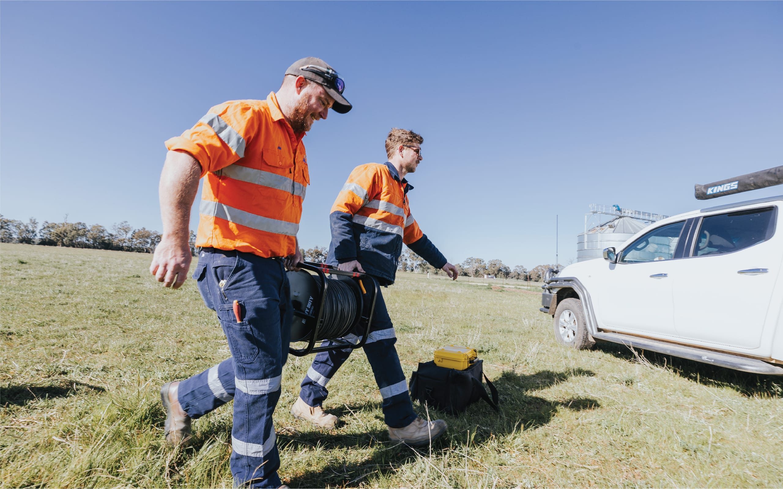 Two MBI Engineering workers in high-visibility uniforms walking across a field beside a white service vehicle