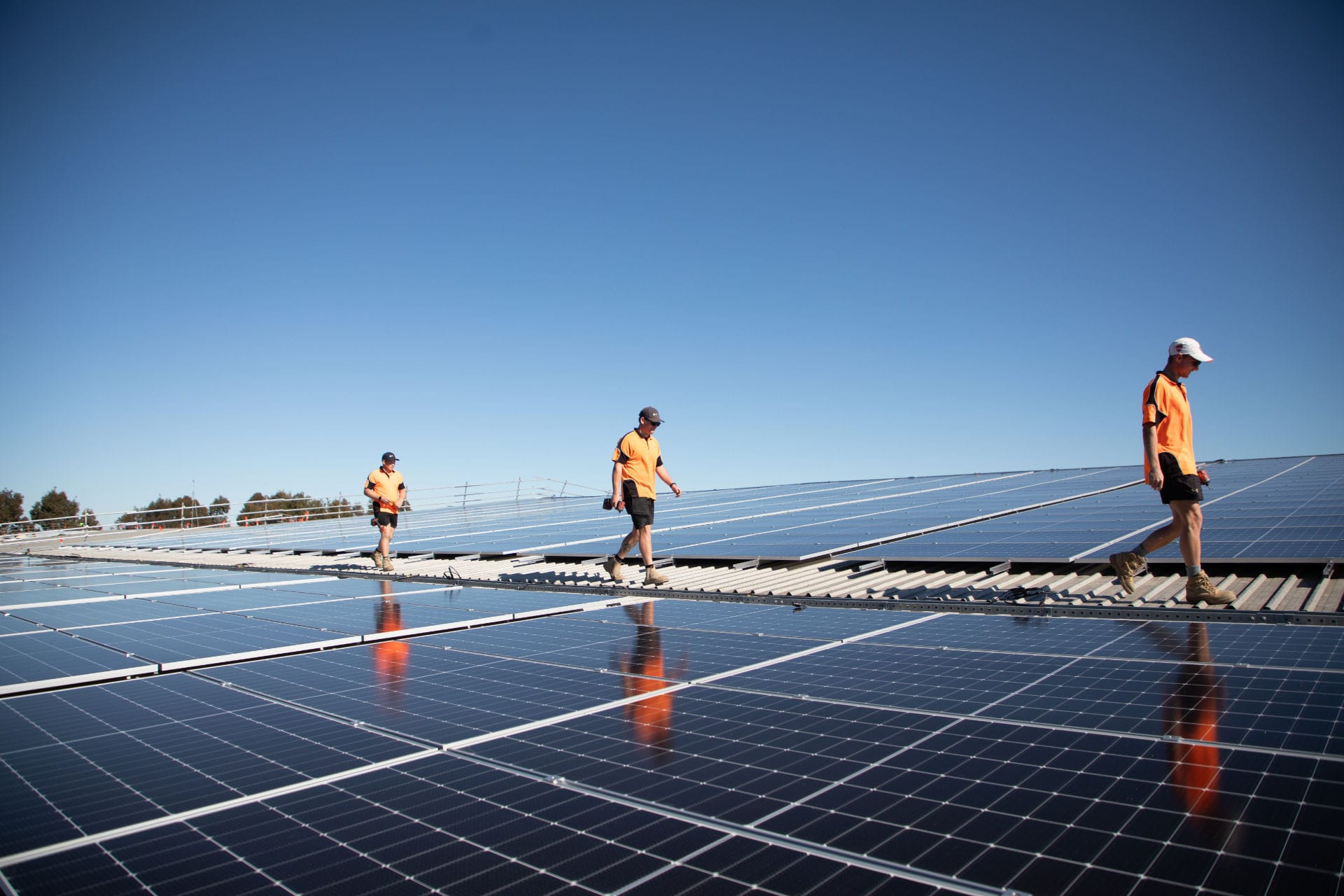 Three solar installers walking across a large rooftop solar panel array under a clear blue sky