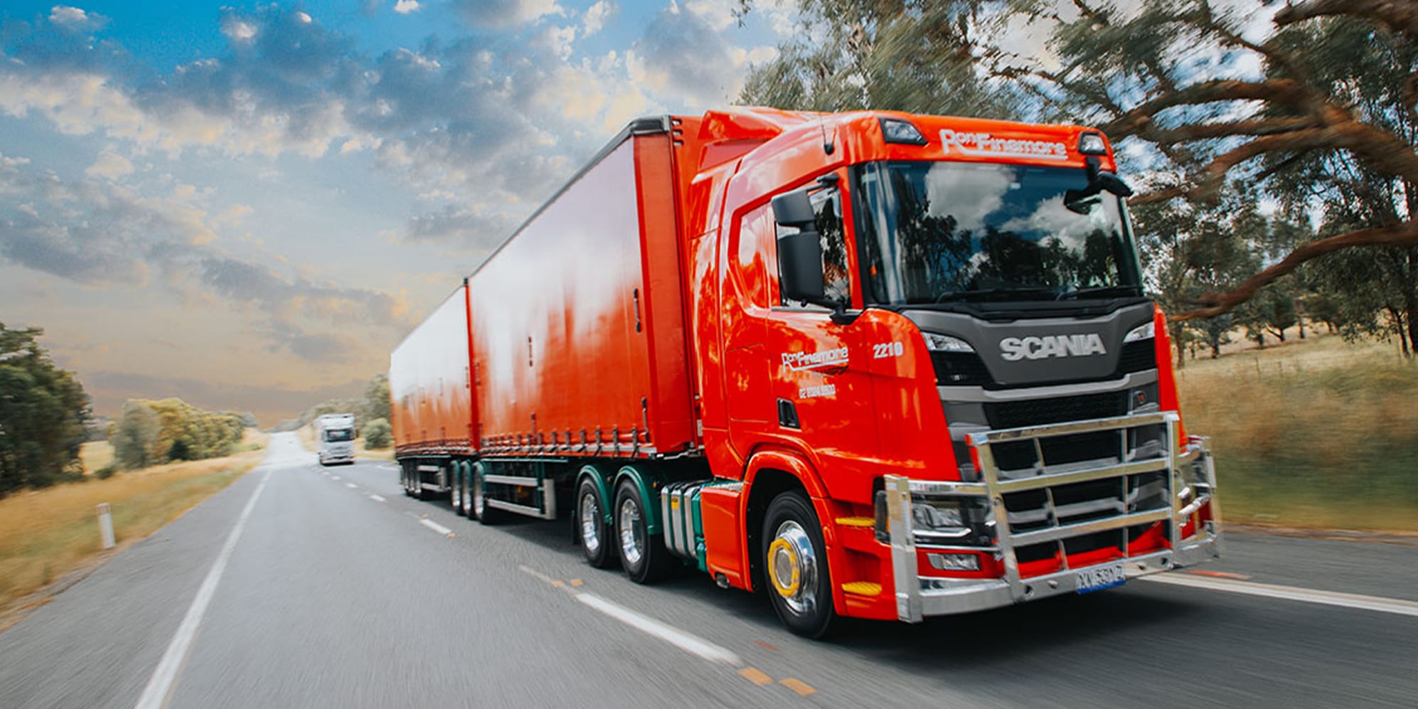 Wide image of a red Ron Finemore Transport Scania truck driving on a country road