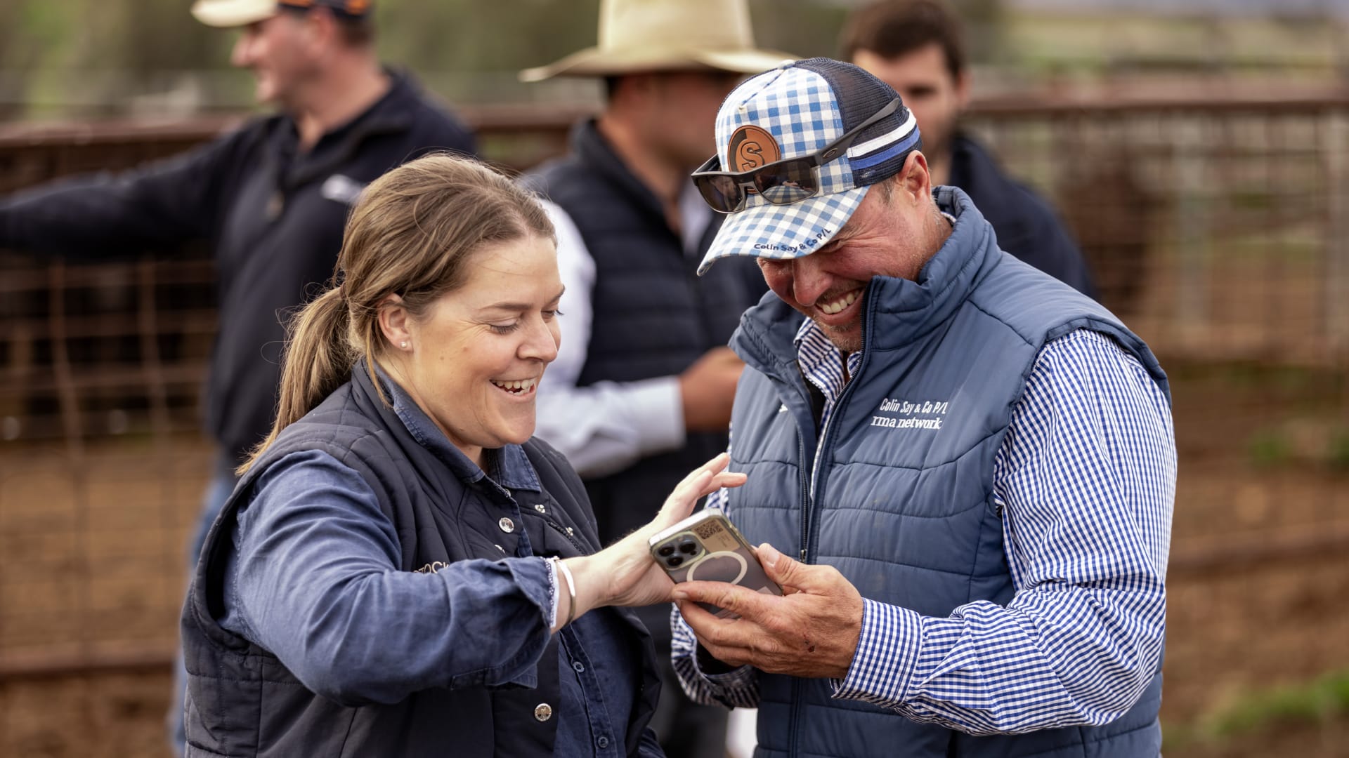 Two livestock workers smiling while reviewing information on a phone at sale yard pens