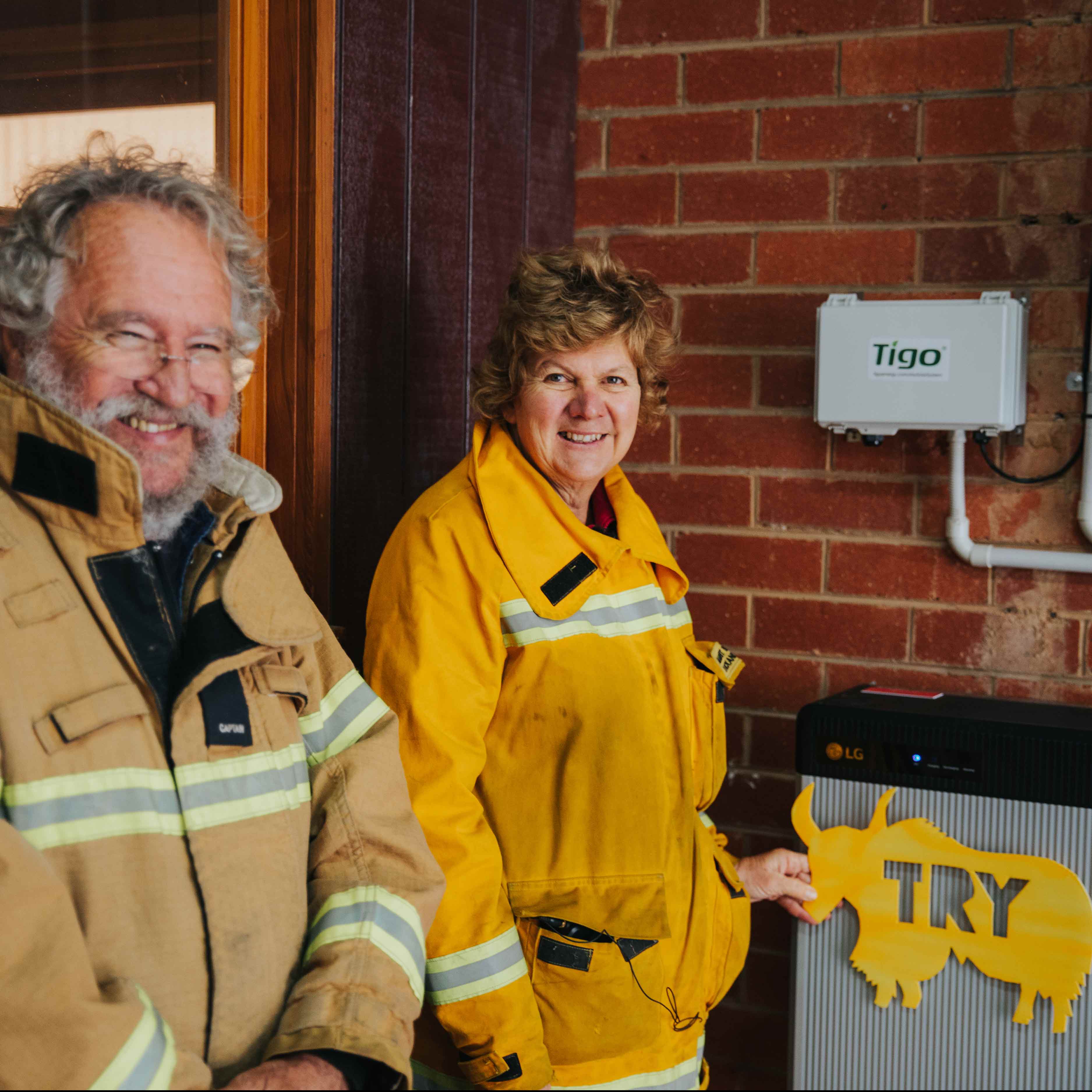 Two people in yellow firefighter jackets standing beside a Tigo solar controller and LG battery
