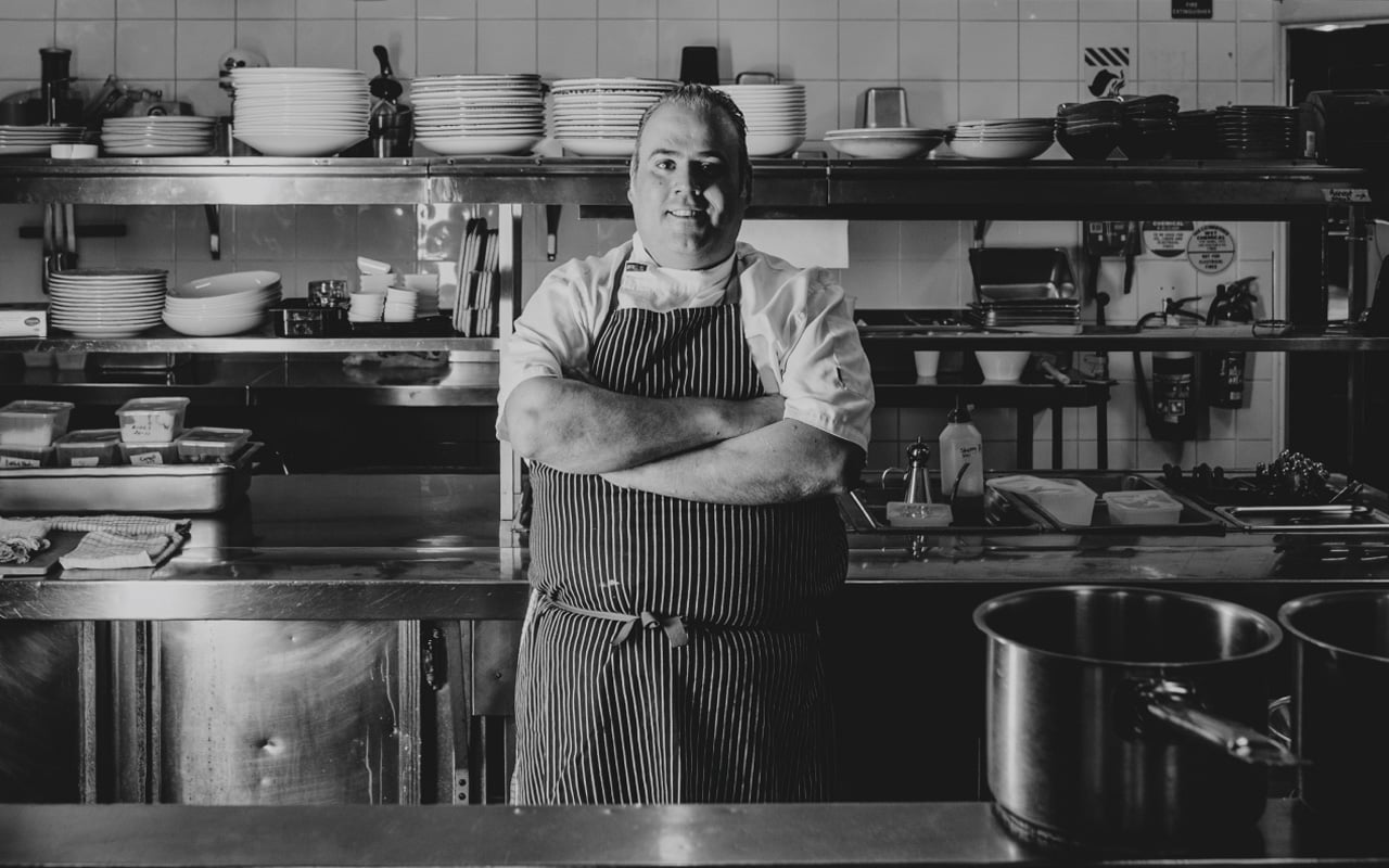 Black-and-white portrait of a chef standing with arms folded in a commercial kitchen