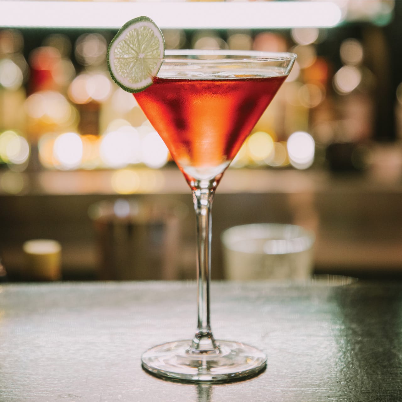 Red cocktail in a martini glass with a lime slice garnish at the bar